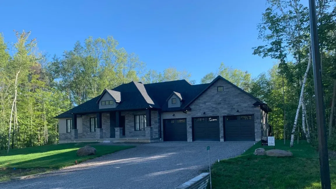 A modern single-story brick house with a dark roof and a three-car garage, surrounded by green trees on a sunny day.
