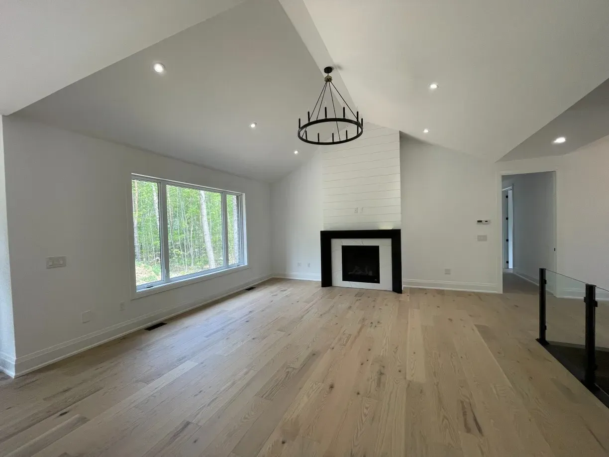 A modern, bright living room with vaulted ceilings, light wood floors, a brick fireplace, and a black chandelier.