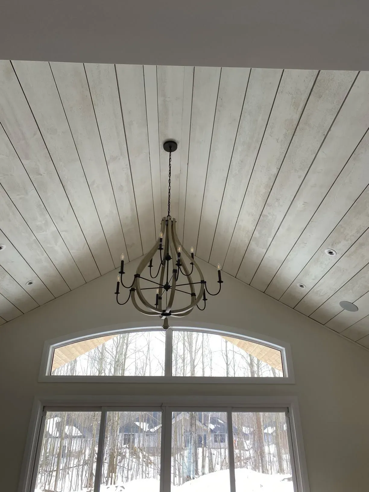 A rustic chandelier hangs from a white-washed, shiplap vaulted ceiling above a large arched window overlooking snowy woods.