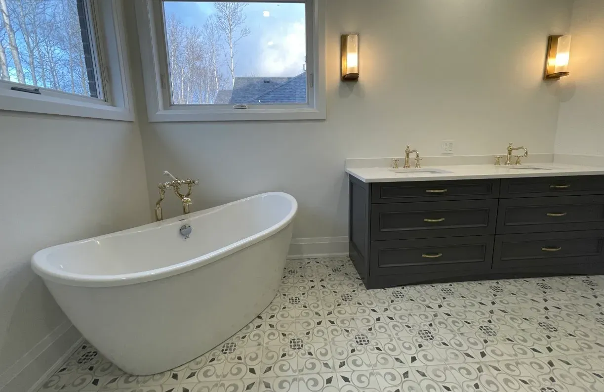 A bright bathroom featuring a white freestanding tub, dark vanity with two sinks, and patterned tile flooring.