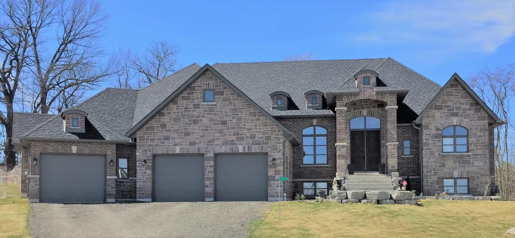 A large, single-story stone house with a dark gray shingled roof and a three-car garage set against a bright blue sky.