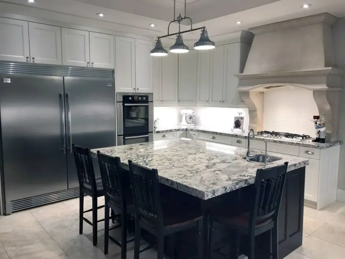 A modern kitchen with white cabinetry, stainless steel appliances, a marble-topped island, and four dark wooden chairs.