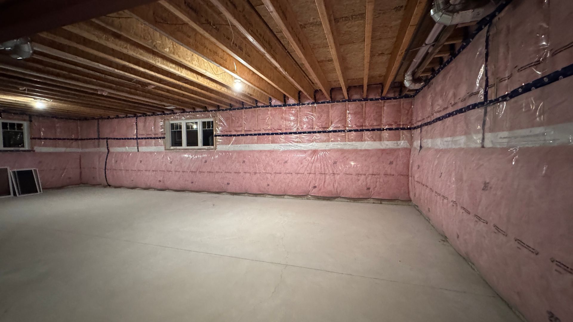 Unfinished basement with pink insulation walls, exposed wooden ceiling, and concrete floor.