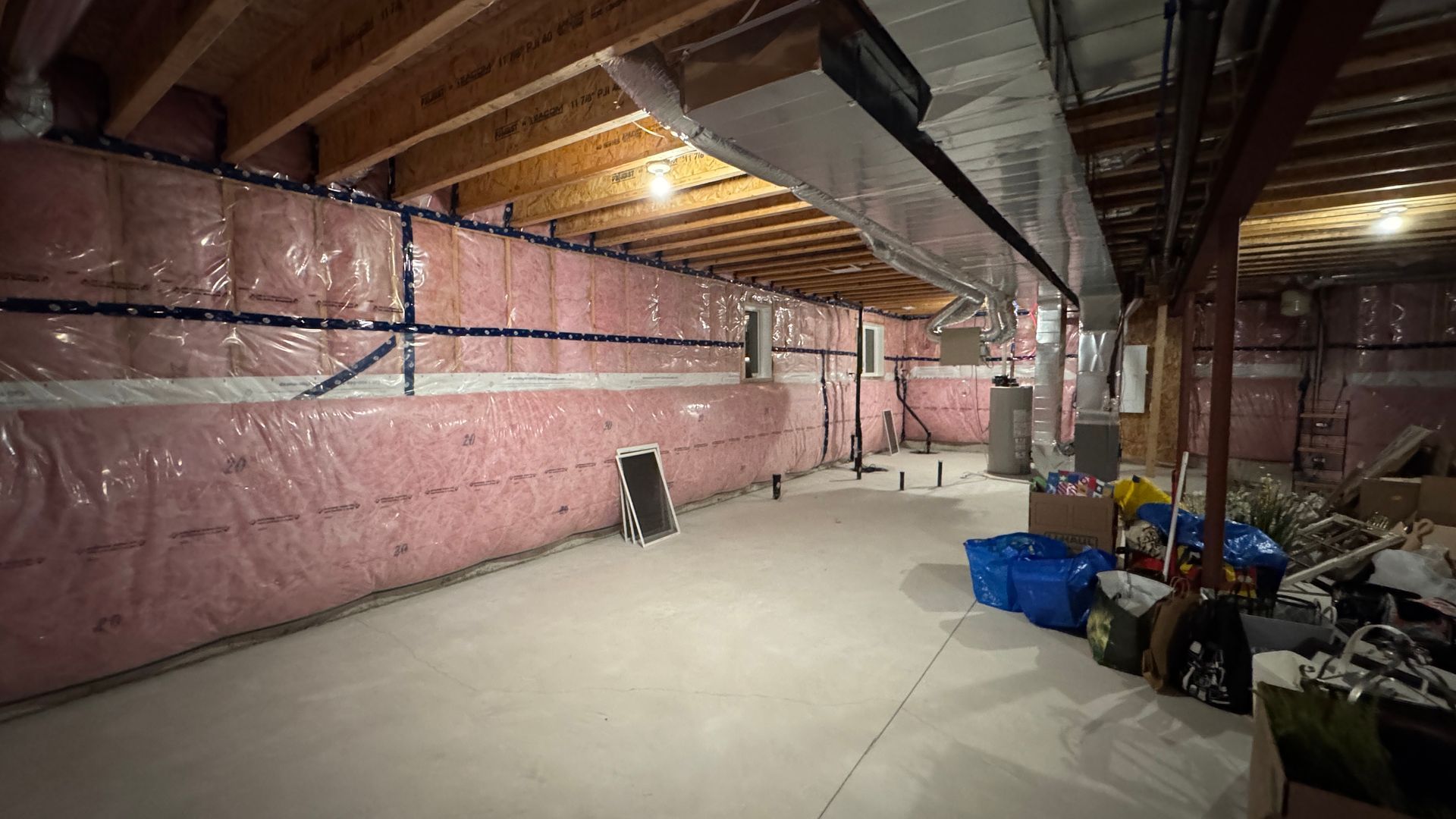 Unfinished basement with pink insulation, exposed beams, and storage bins along a concrete floor.