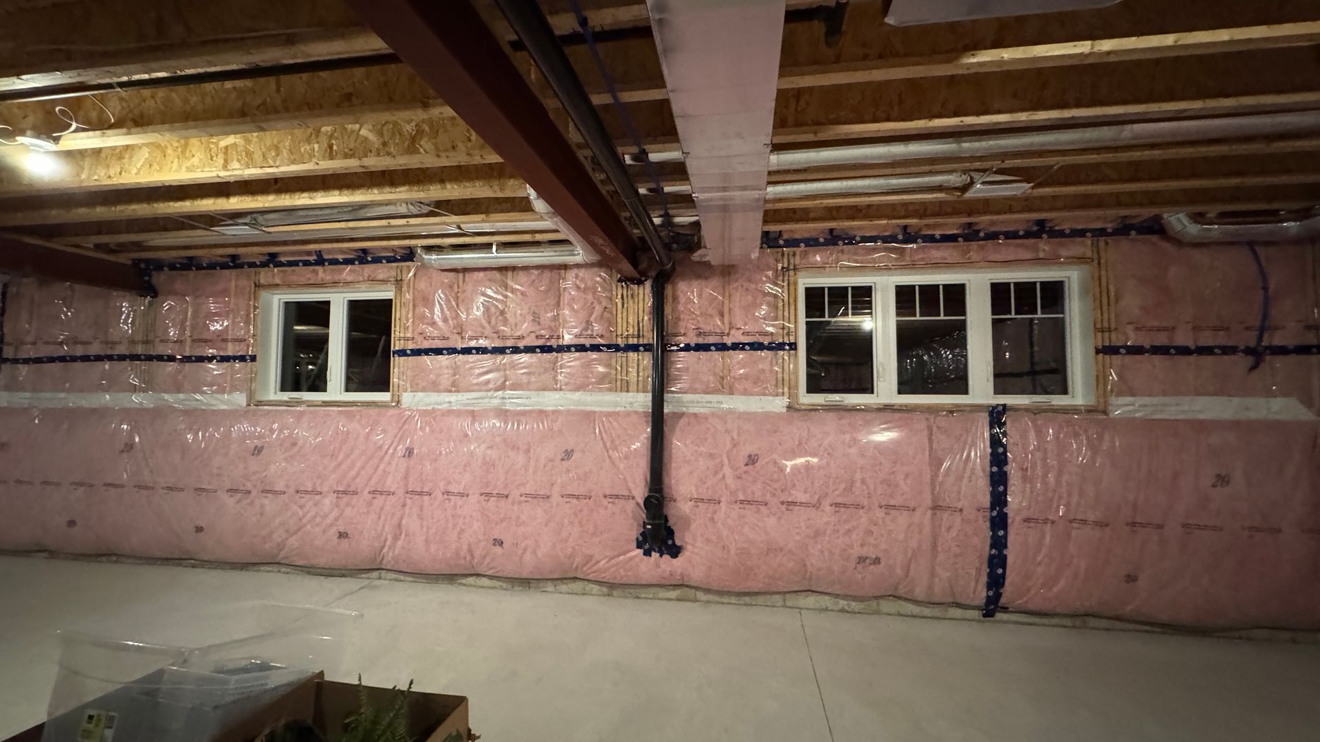 Basement wall with pink insulation, exposed beams, two windows, and a central support post