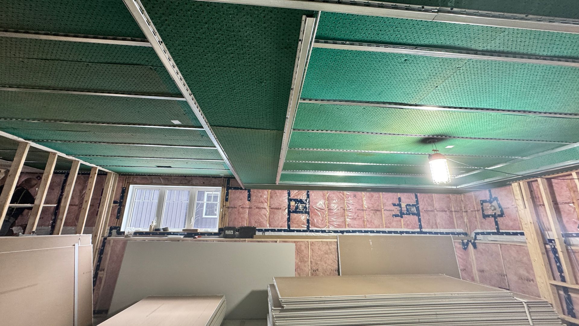 Unfinished basement room with exposed ceiling, green insulation, windows, and mattresses on the floor