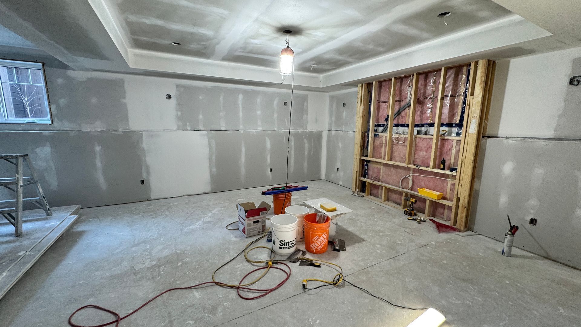 Renovation room with unfinished drywall, exposed studs, and paint buckets on a bare floor.