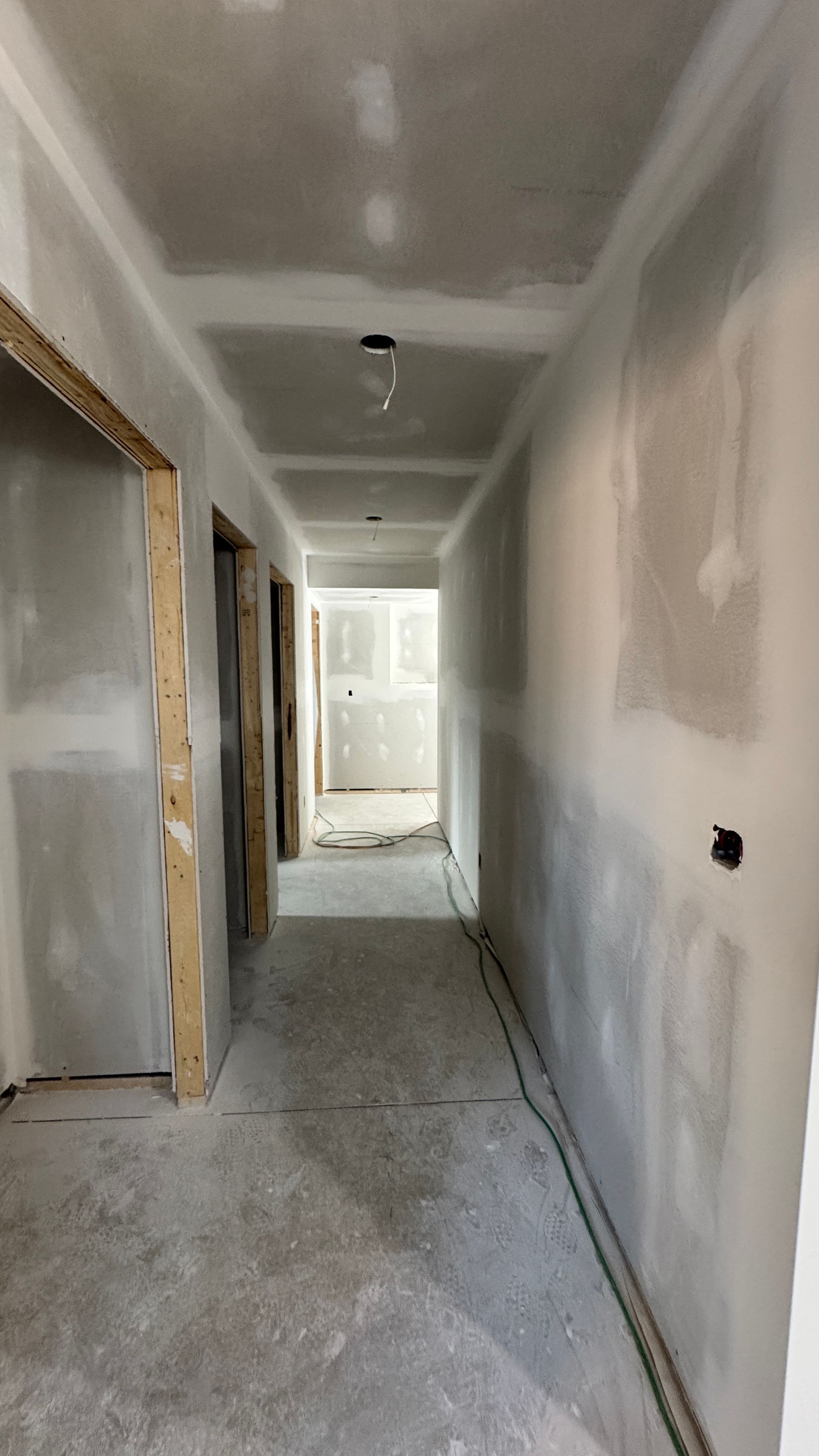 Narrow unfinished hallway with drywall, exposed doorways, and bright light at the far end