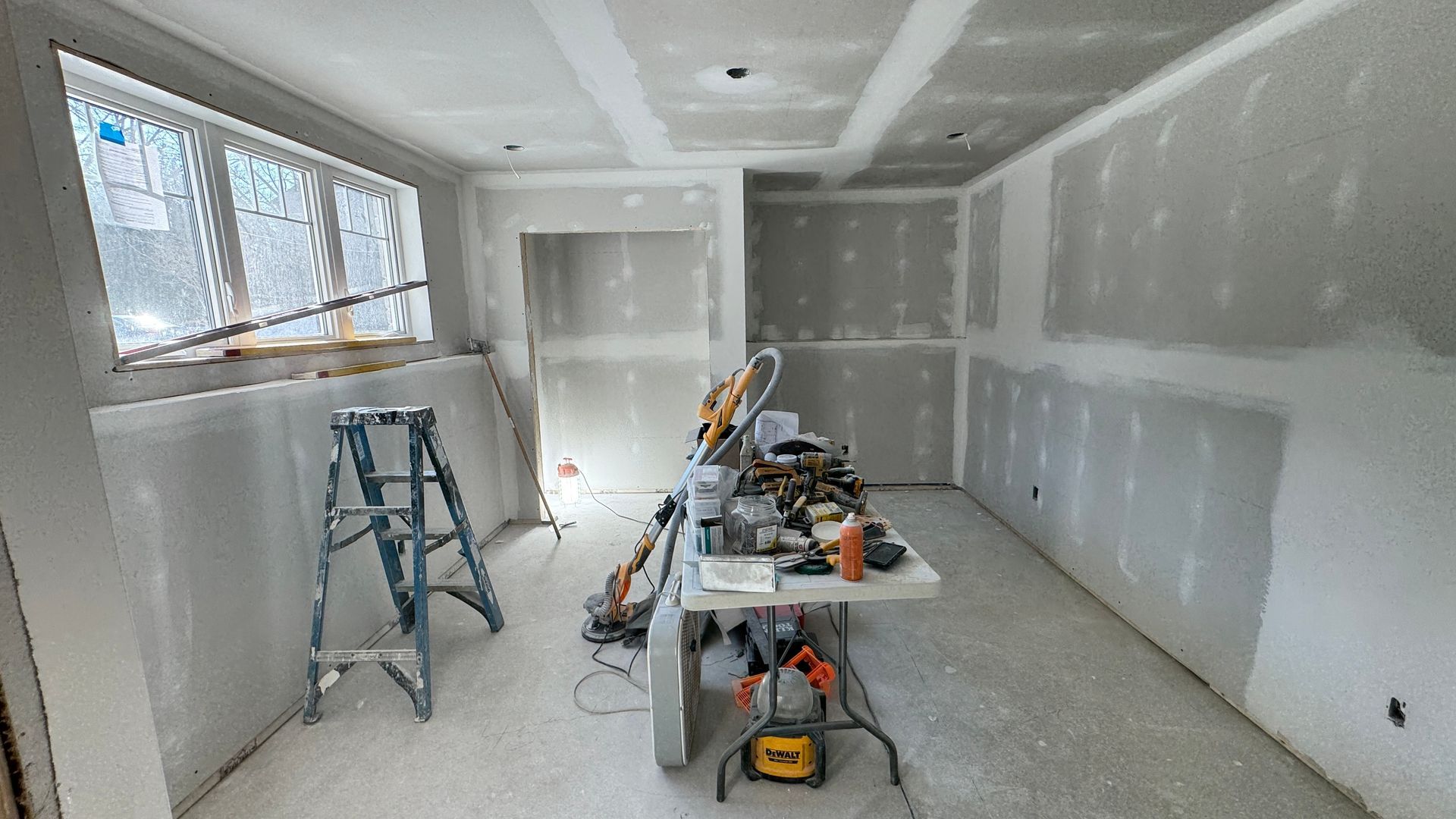 Partially renovated room with drywall, ladders, tools, and a worktable in the center.