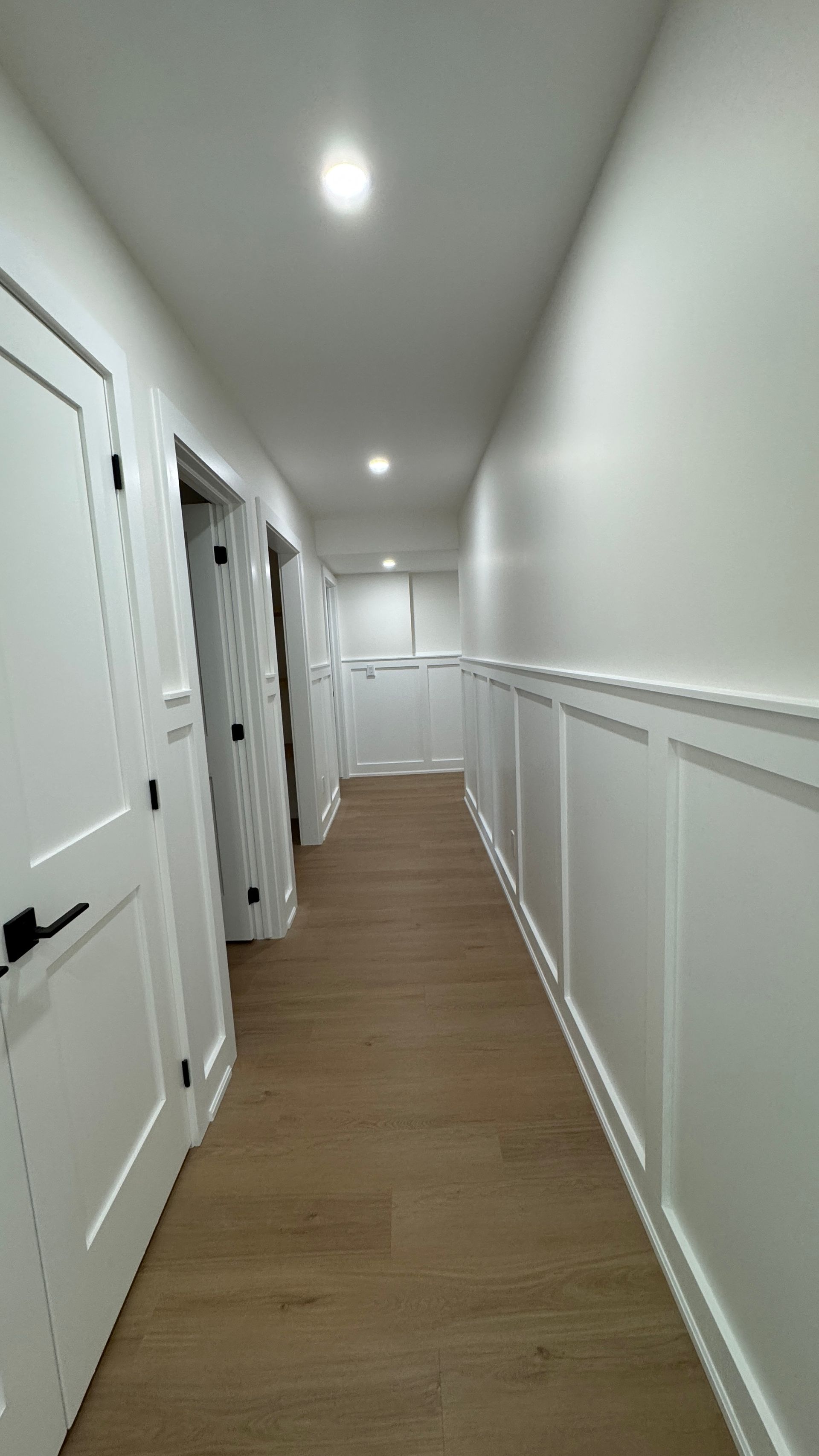 Narrow white hallway with wood floors, recessed ceiling lights, and paneled walls