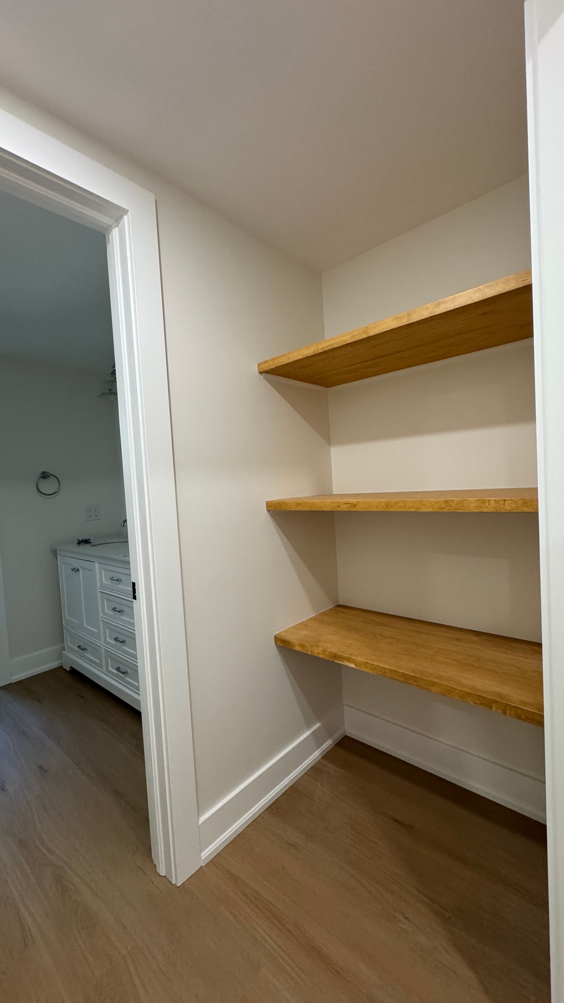 Small closet with three wooden shelves and white walls beside an open doorway, on wood flooring.
