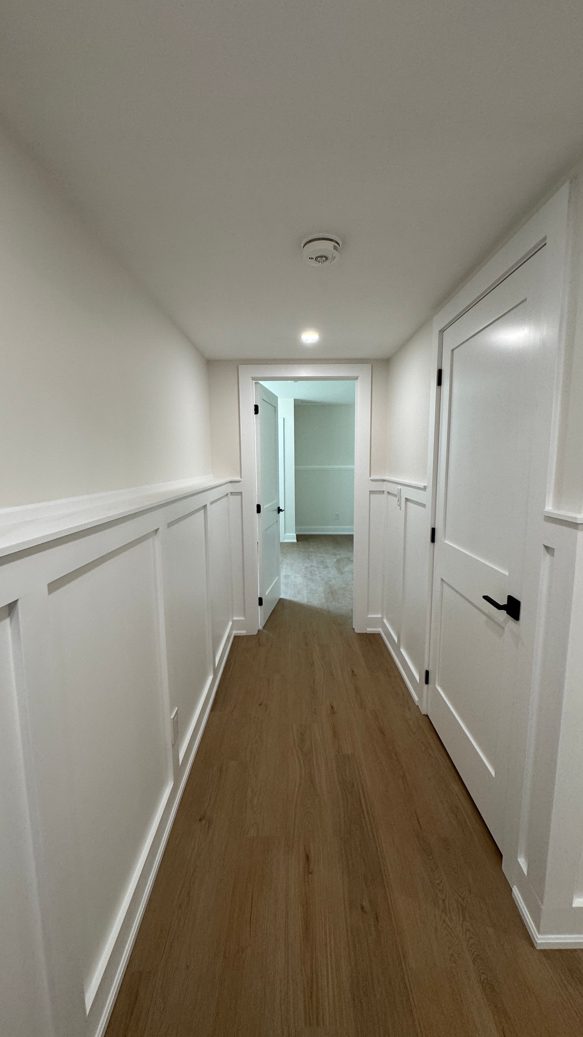 Narrow white hallway with wood flooring, doors on both sides, and a lit doorway at the end