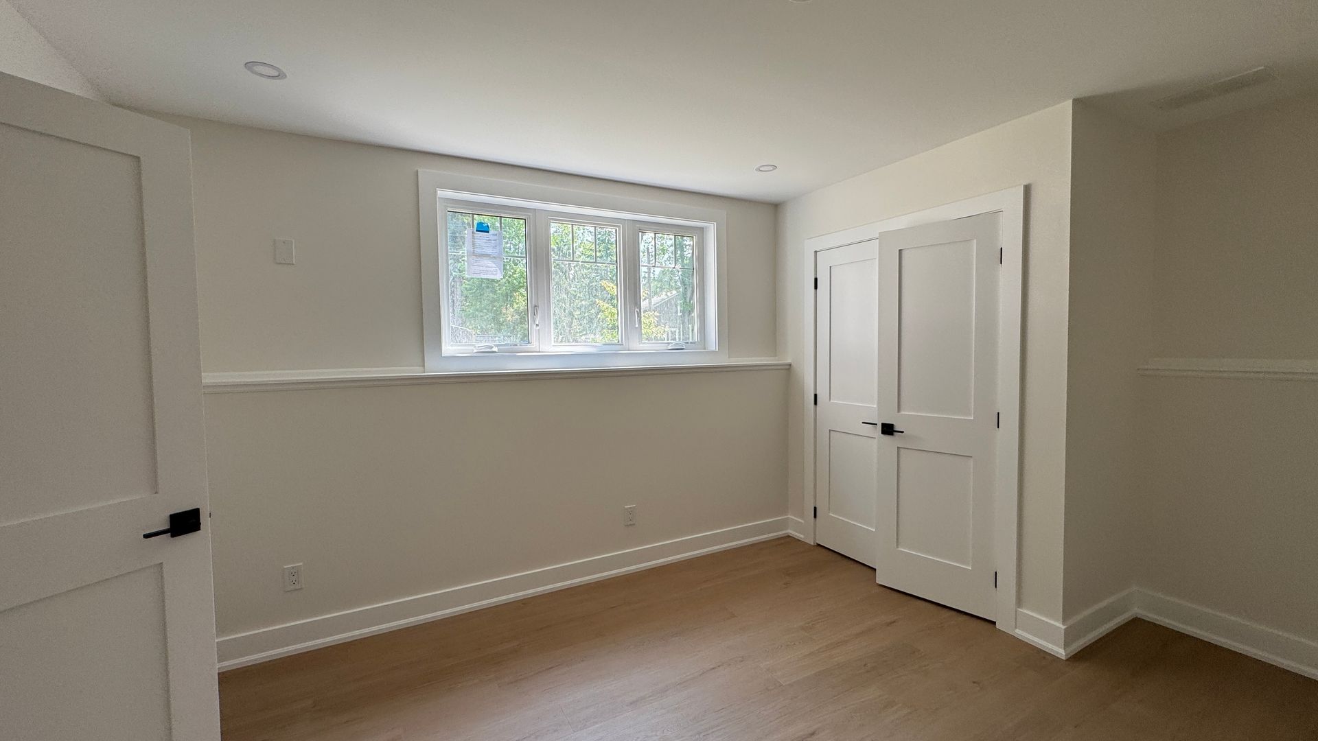 Empty white bedroom with a small window and double closet doors, light wood floor.