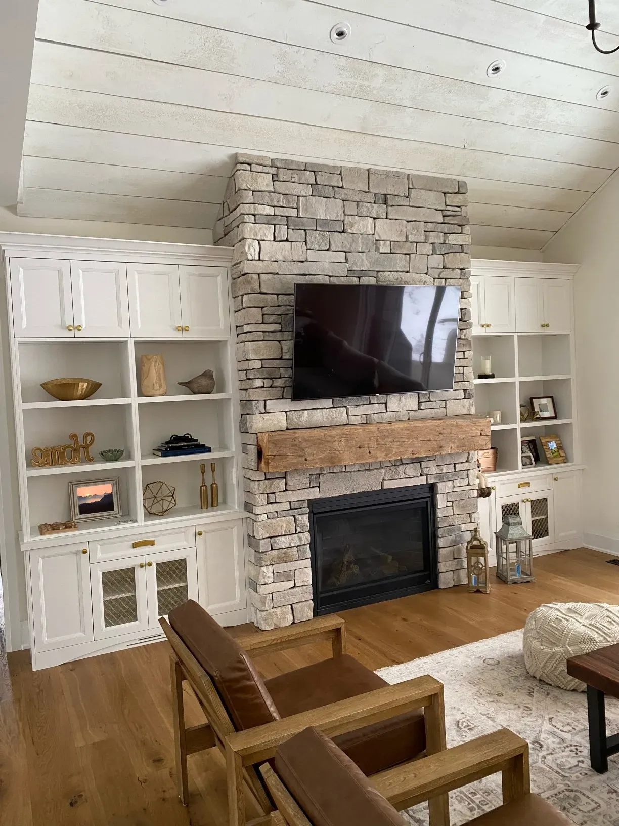 A white living room features a tall, stacked stone fireplace with a rustic wooden mantel, flanked by white built-in shelves.