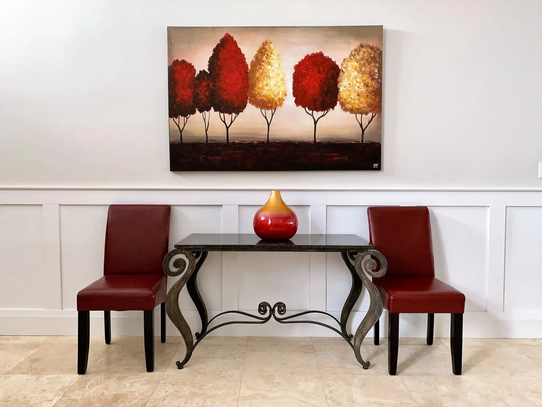 A table with two red chairs and a decorative vase sits against a wall beneath a painting of stylized red and gold trees.