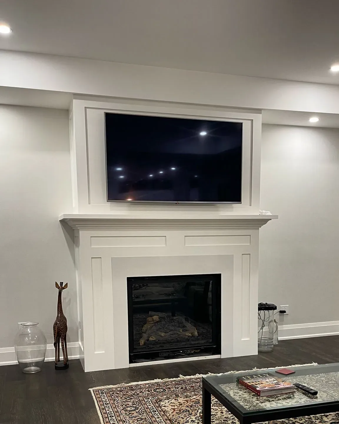 A white fireplace mantel with a mounted television above it, flanked by a glass vase, a giraffe sculpture, and a side table.