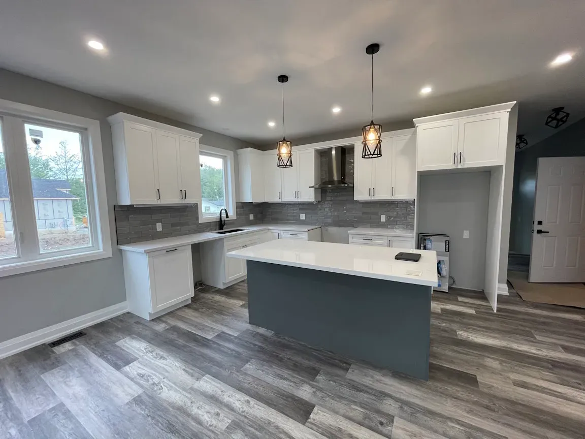Modern kitchen with white upper cabinets, dark island, gray backsplash, and light wood-patterned flooring.