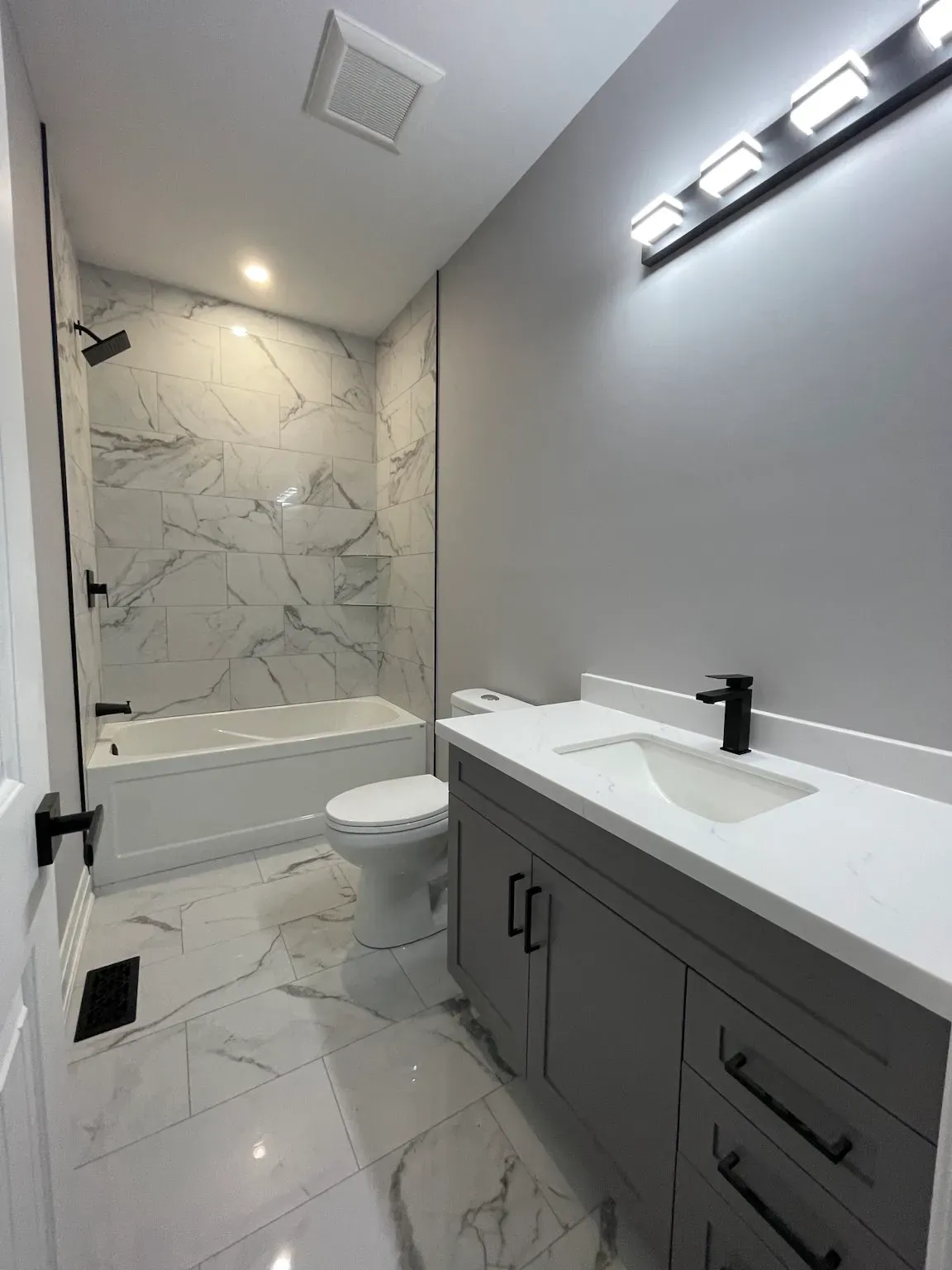 A modern bathroom with a gray vanity, white quartz countertop, toilet, and a bathtub with white marble-patterned tiles.