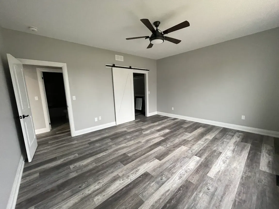 A spacious room with grey walls, a ceiling fan, wood-look flooring, a standard doorway, and a sliding barn-style door.