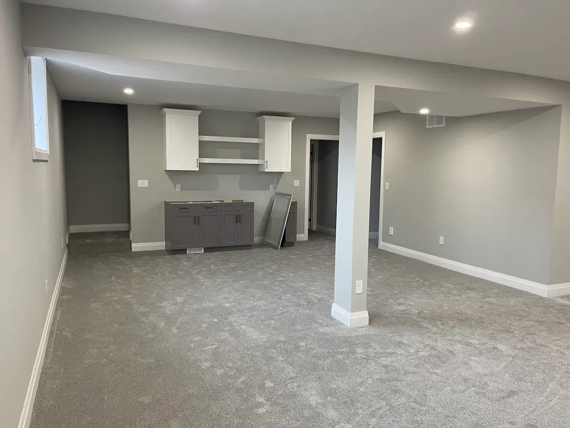 A carpeted basement with gray walls, a support pillar, recessed lighting, and a small kitchen area with gray cabinets.