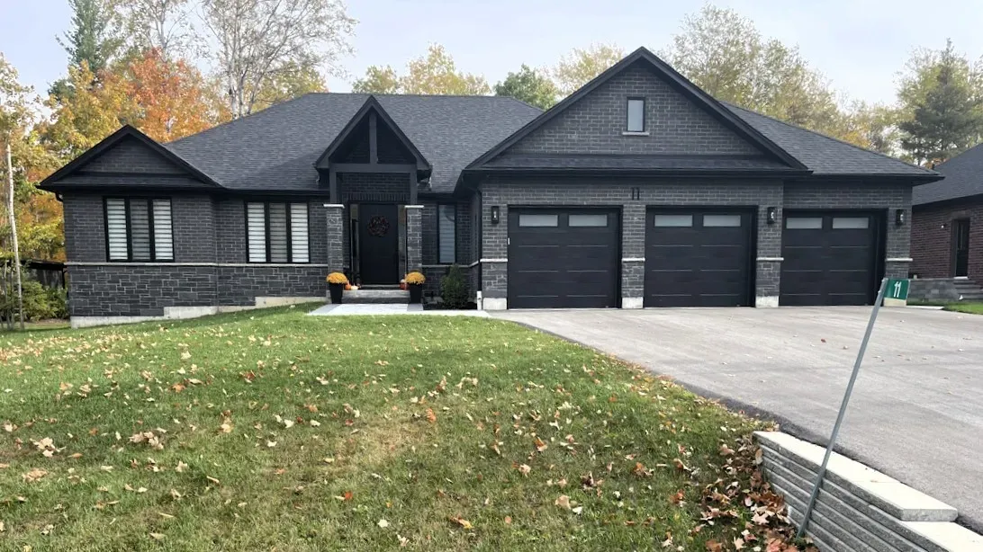 A dark-toned, single-story suburban house with a three-car garage, stone facade, and a lawn covered in fallen leaves.