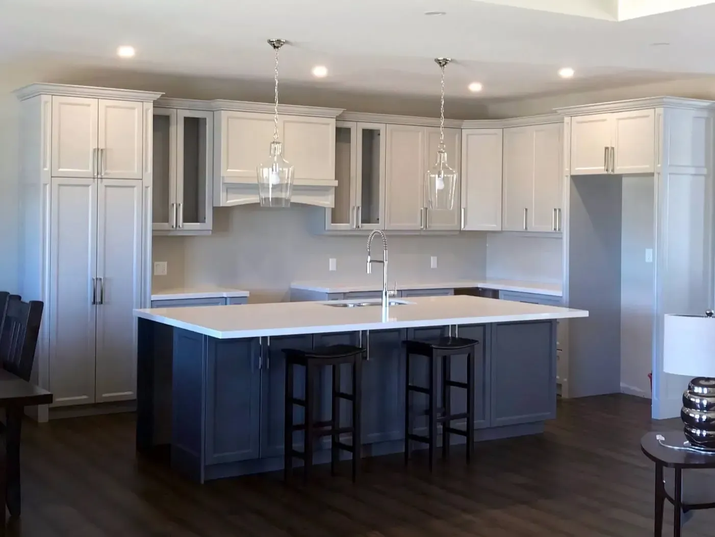 A kitchen featuring white wall cabinets, a large dark gray island with two stools, white countertops, and wood flooring.
