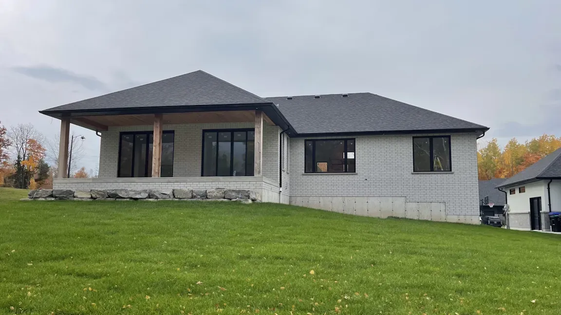 A modern, unfinished concrete house with a dark gabled roof and a covered porch stands on a grassy slope in autumn.