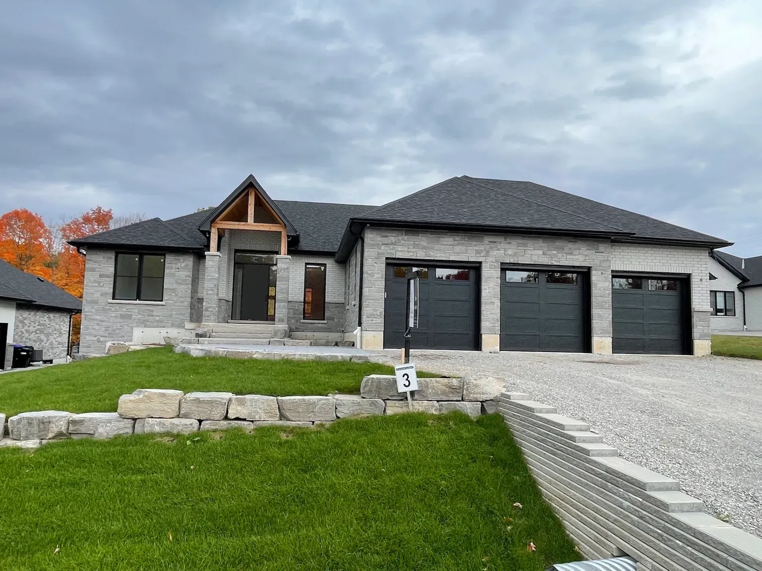A newly constructed single-story stone house with a three-car garage and a front porch, set on a sloped, grassy lot.