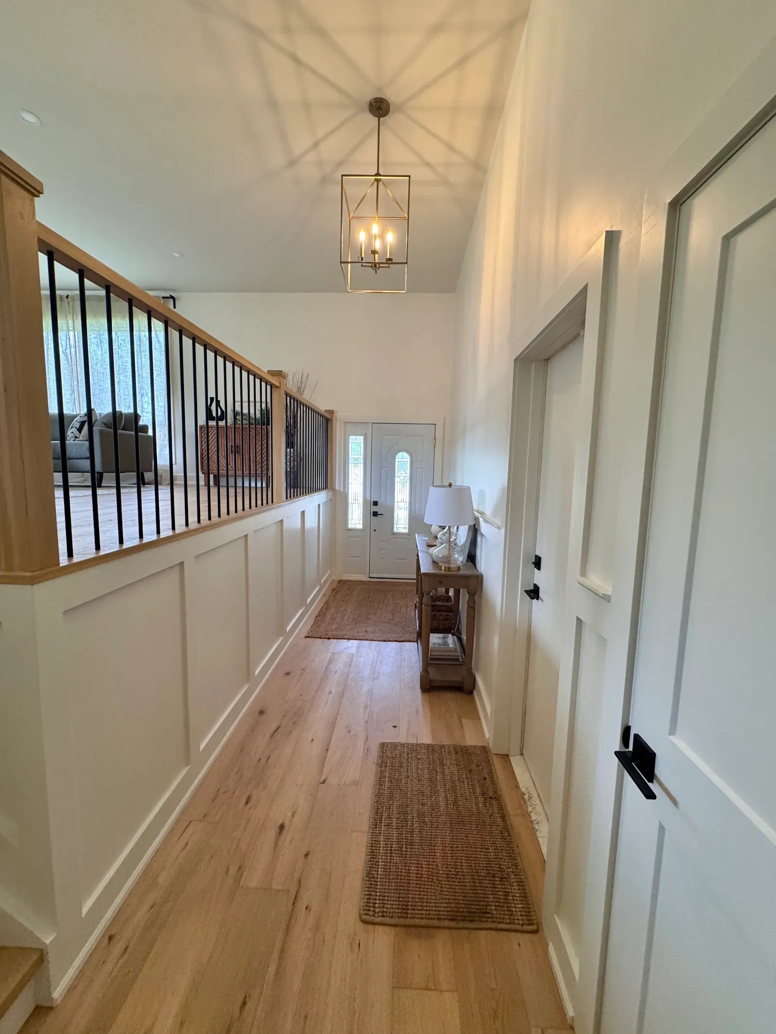 A hallway with light wood floors, white board-and-batten walls, a metal railing, and a modern lantern pendant light.