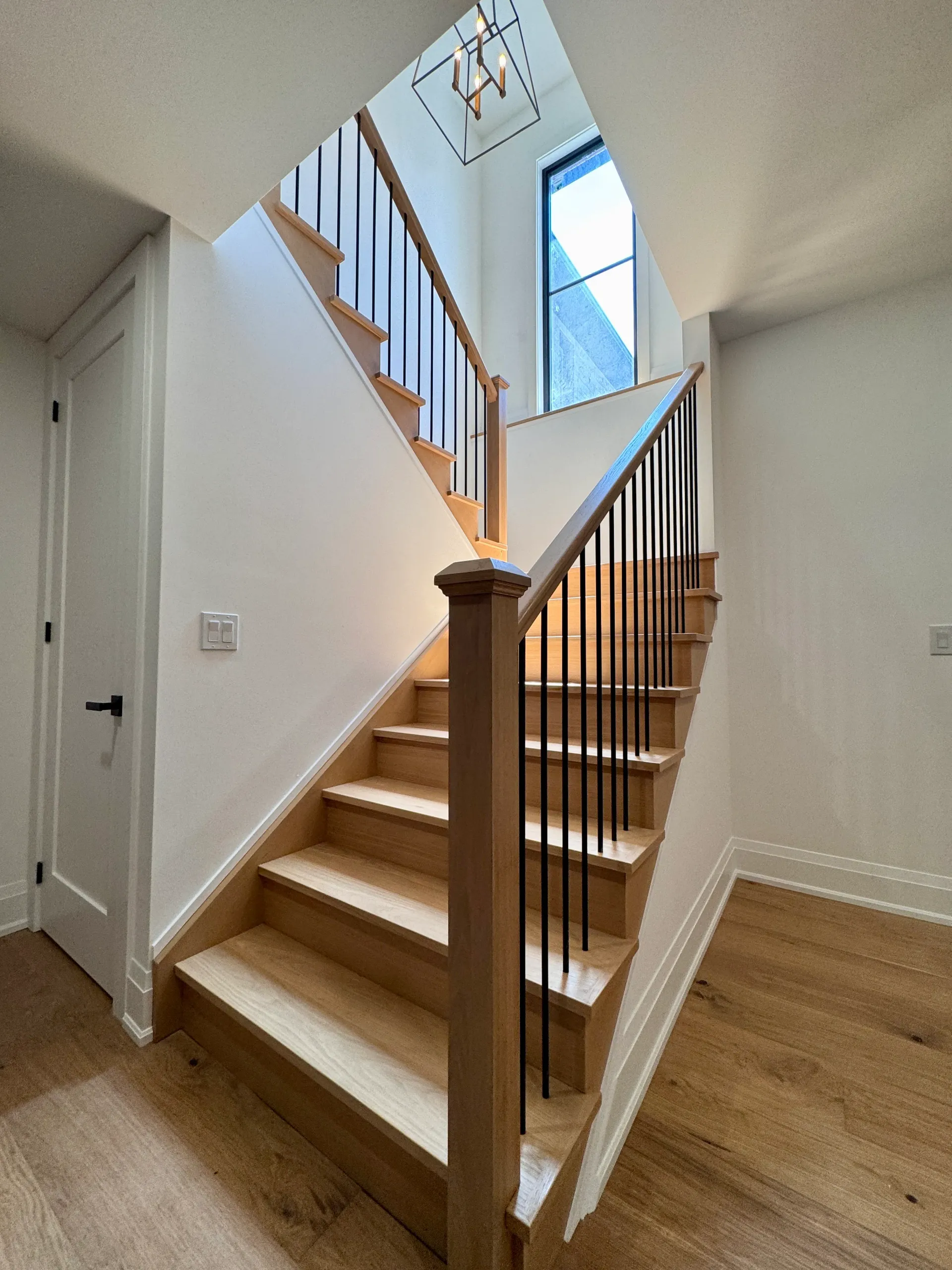 A wooden staircase with black metal balusters leads upward toward a bright window and a modern pendant light.