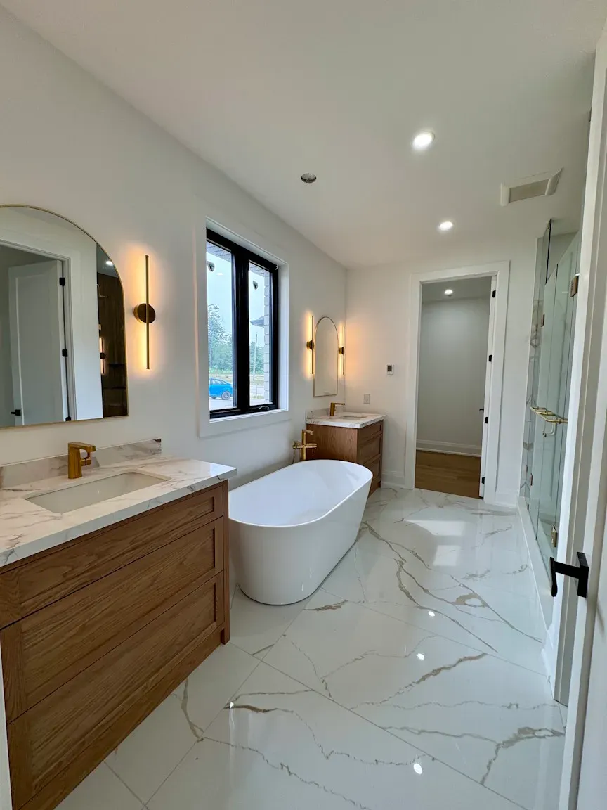 A modern bathroom with marble floors, a white freestanding tub, a wood vanity, and a black-framed window.