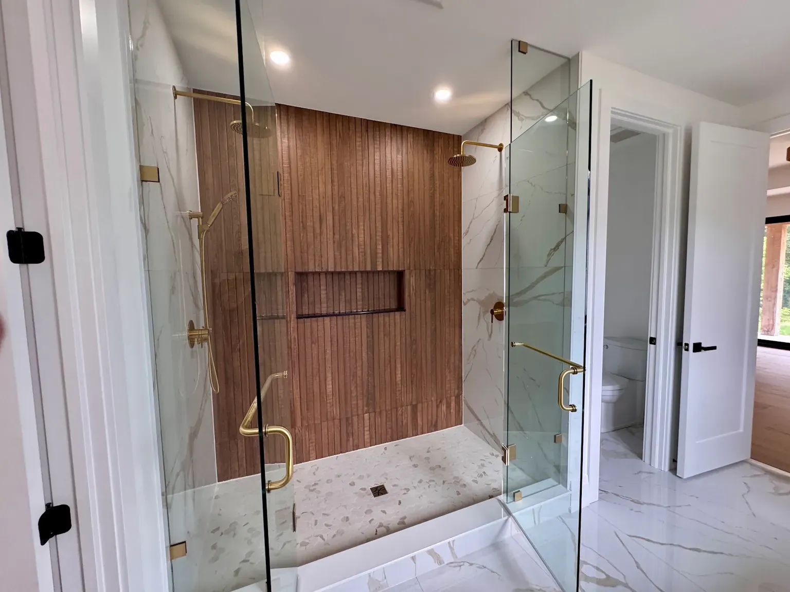 Modern bathroom shower with glass doors, a wood-paneled accent wall, a built-in niche, and white marble-style flooring.