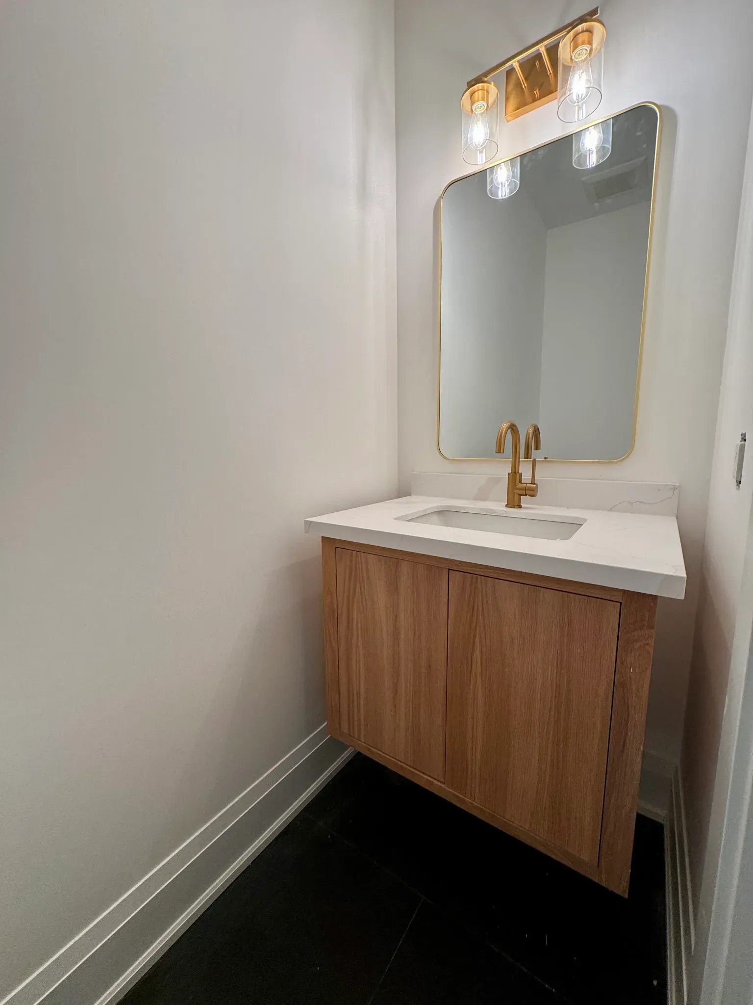 A modern bathroom vanity with a wood cabinet, white countertop, gold faucet, and an arched mirror on a white wall.