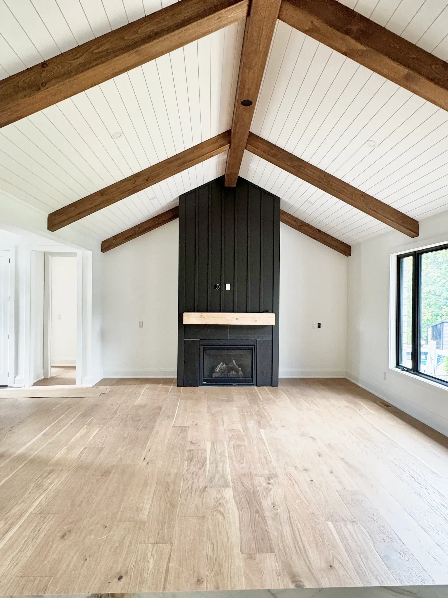A room with vaulted white ceilings, exposed wood beams, light wood floors, and a dark, vertical wood-paneled fireplace.