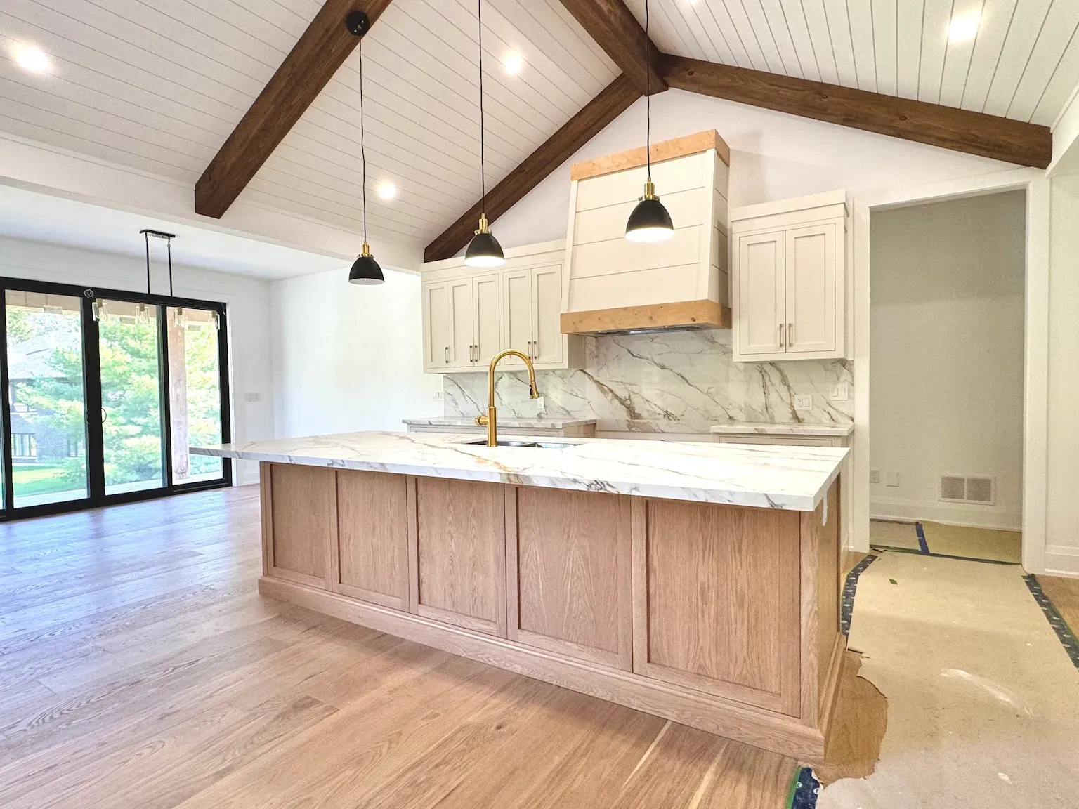 A modern kitchen with light wood cabinets, white marble countertops, a farmhouse-style vent hood, and vaulted ceilings.