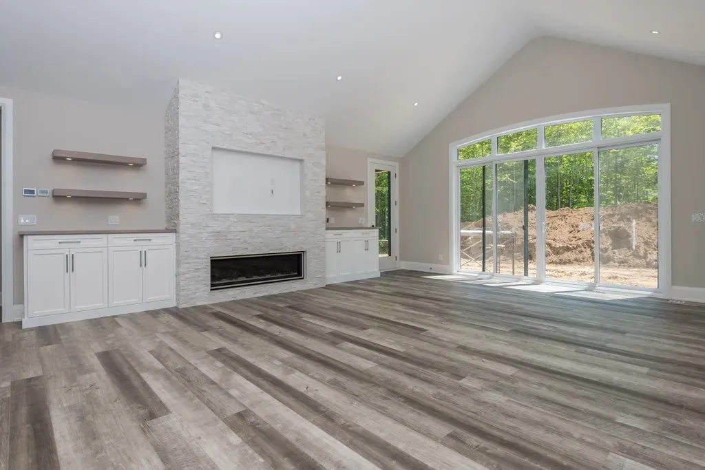 Modern living room with wood-look flooring, a white stone fireplace wall, and large sliding glass doors overlooking woods.