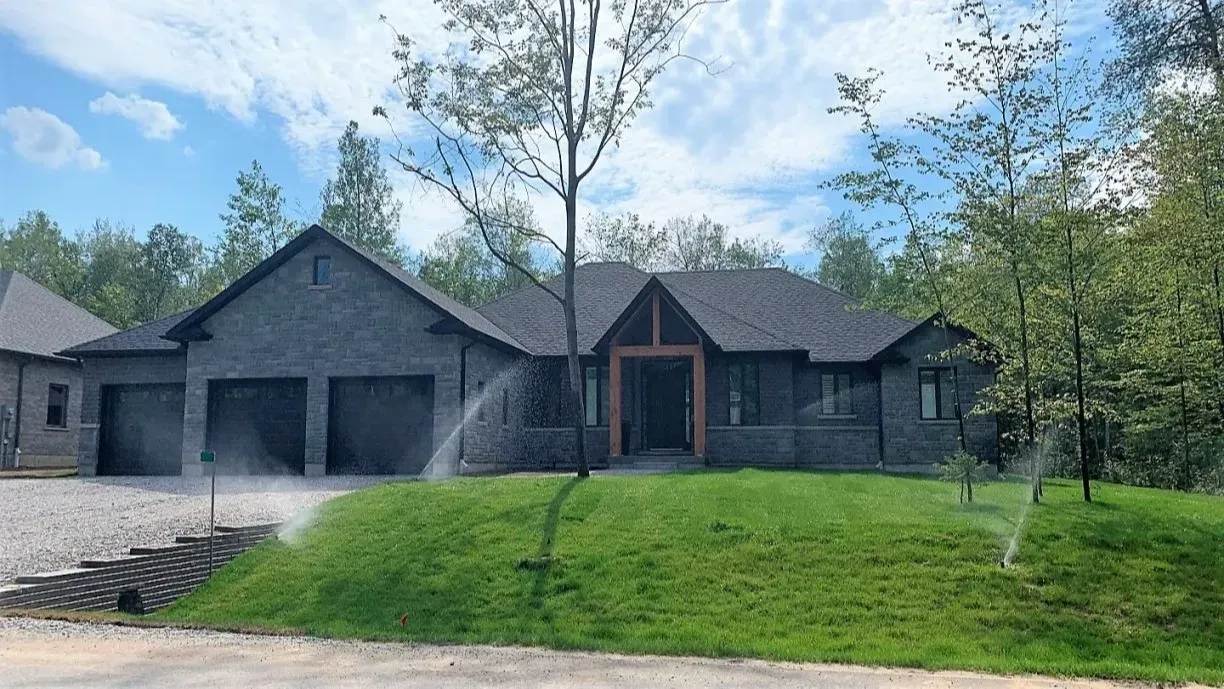 A modern grey brick house with a dark roof and a central wooden entryway, surrounded by trees with lawn sprinklers active.