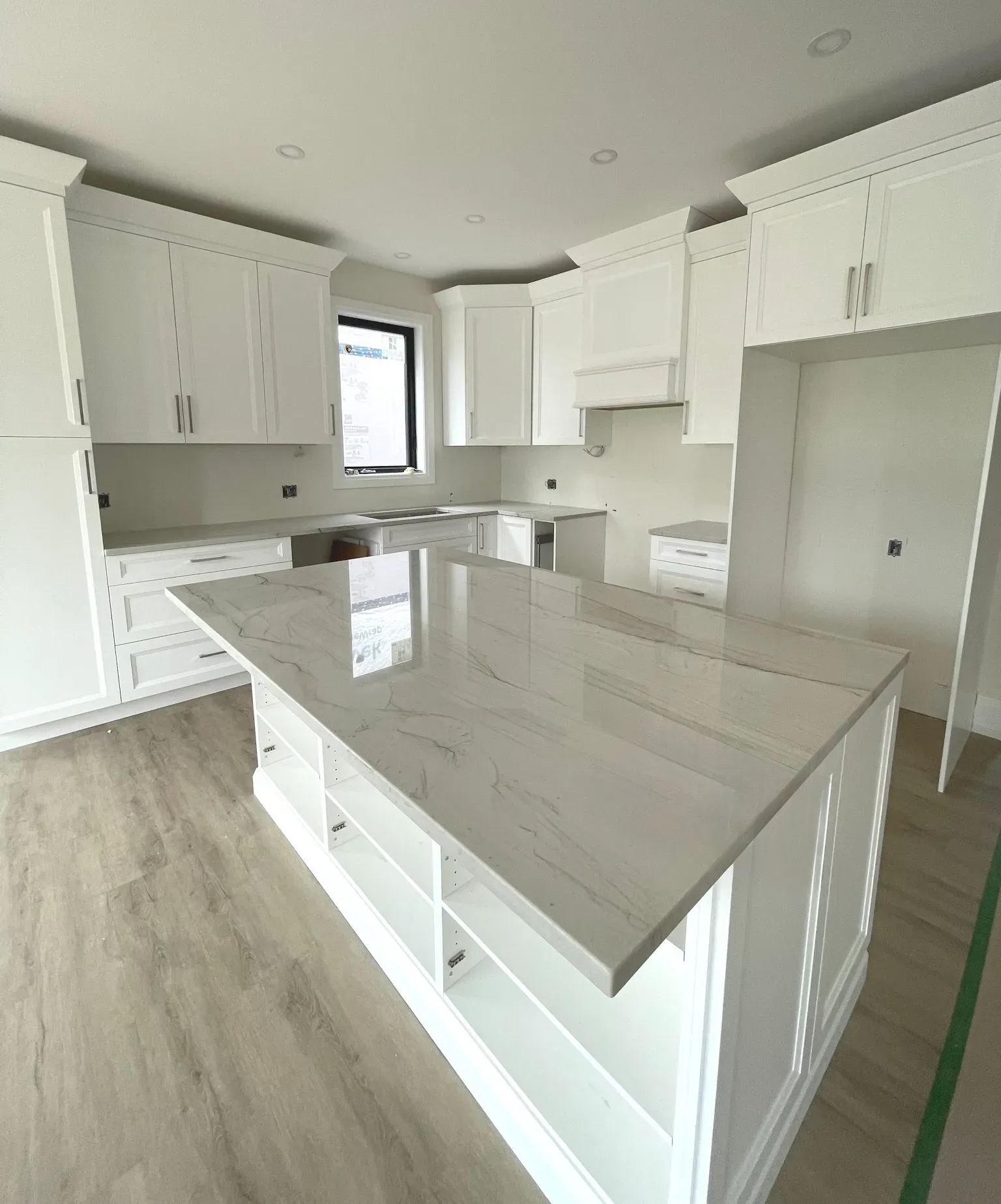 A white kitchen with a large marble-topped island, wooden floors, and new cabinetry, illuminated by recessed lighting.