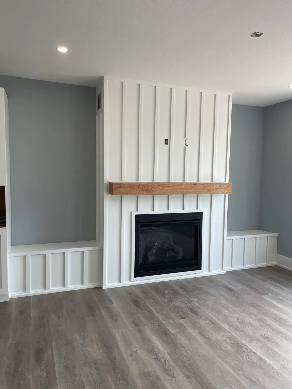 A modern living room features a fireplace with a white board-and-batten accent wall, wood mantel, and built-in side benches.