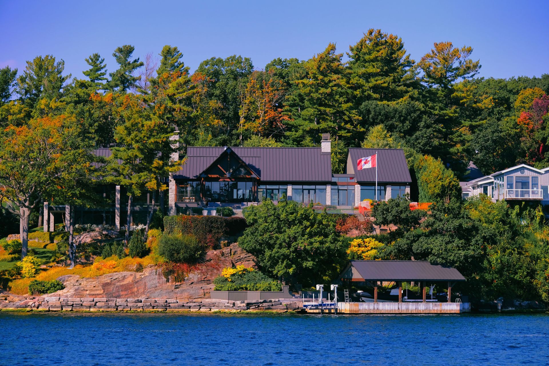 Lakeside homes with autumn trees, rocky shore, and blue water under a clear sky.