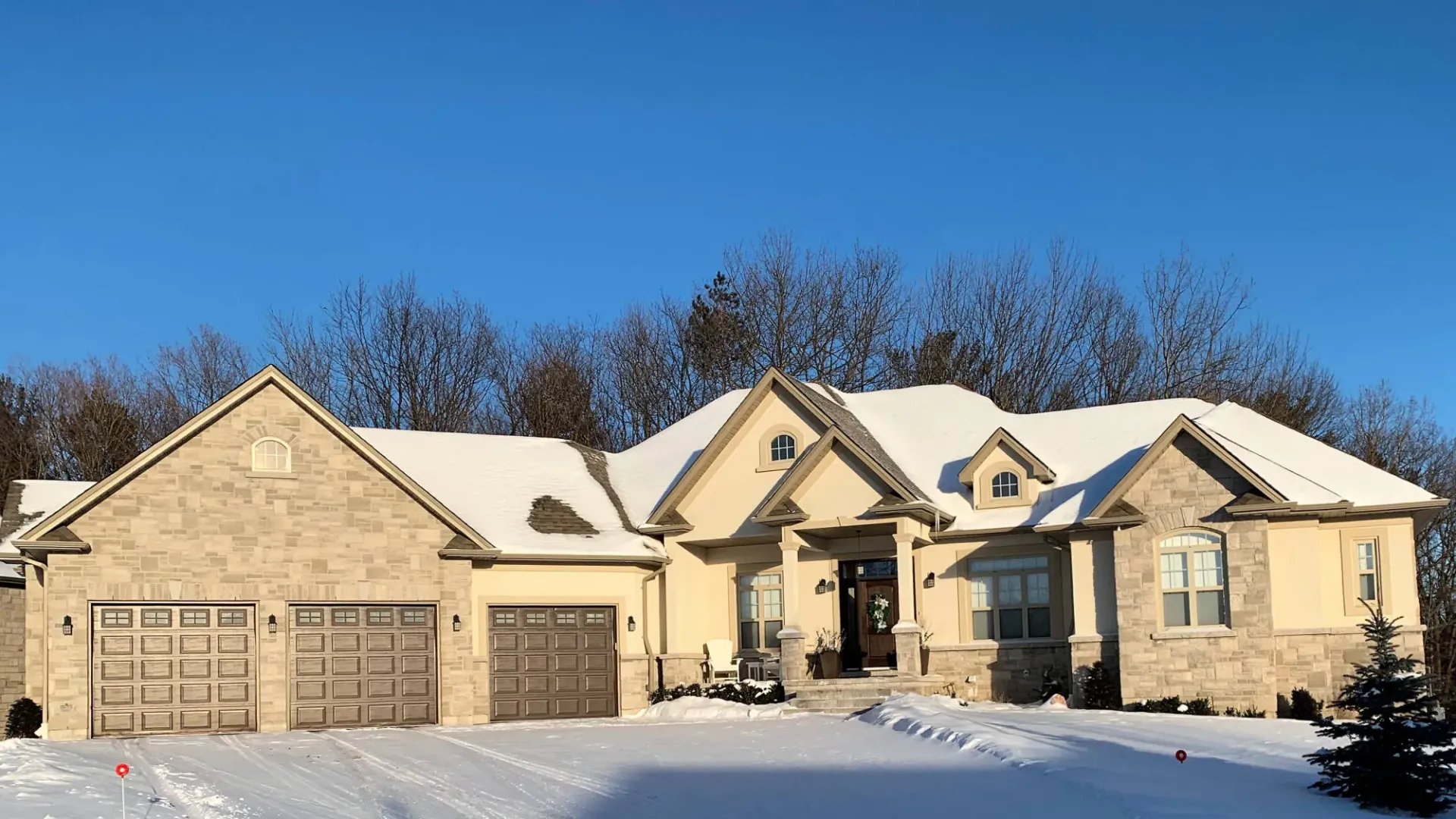 A tan stone house with a three-car garage and snow-covered roof against a clear blue sky.