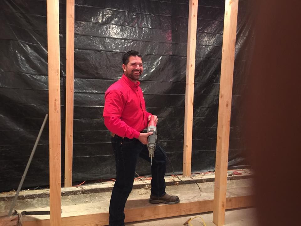 Man in red shirt, holding drill, standing inside a wooden frame. Smiling, working in a construction setting.