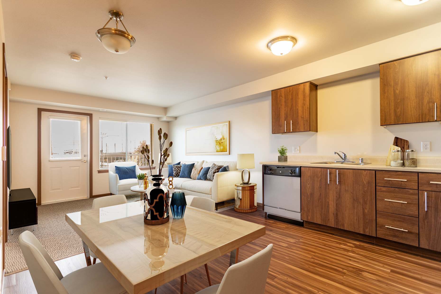 Open-concept apartment with kitchen, dining area, and living room. Light wood cabinets, white walls, beige and blue furniture.