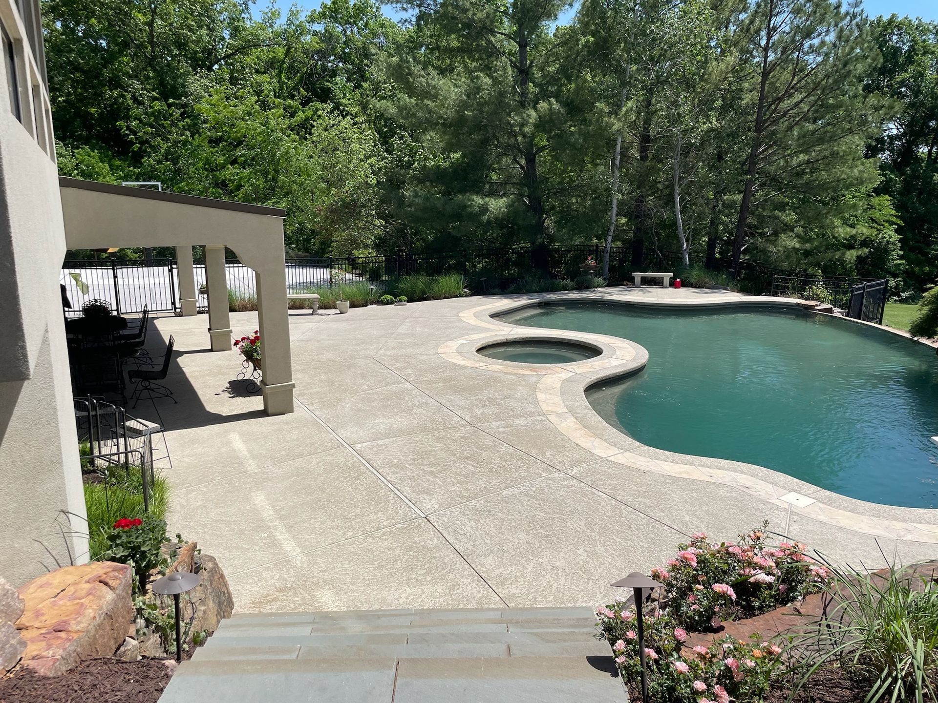 Curved backyard swimming pool beside a white patio and trees, with a stone deck and flower beds.