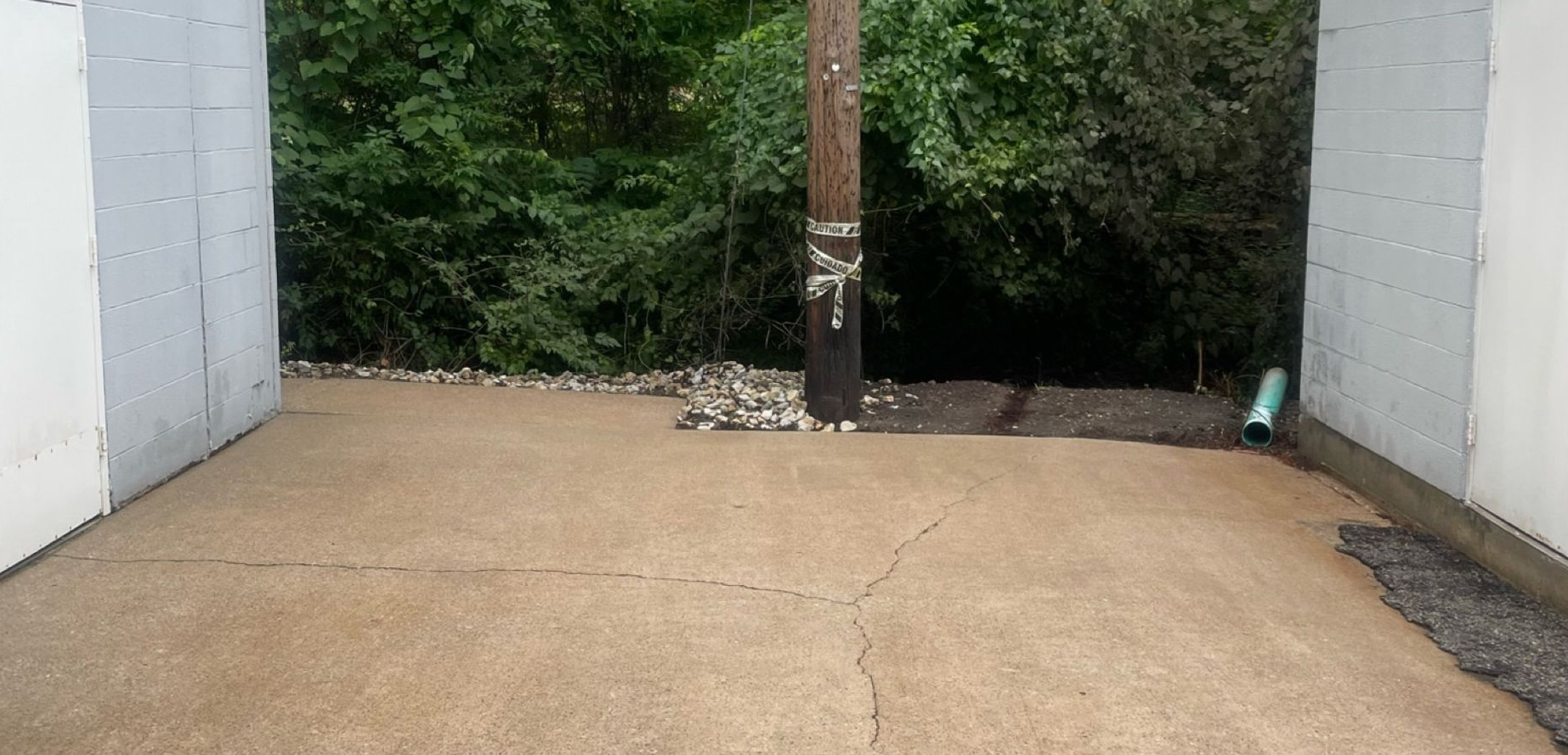 Concrete driveway between two white walls leading to a wooded area, with a utility pole in the center.