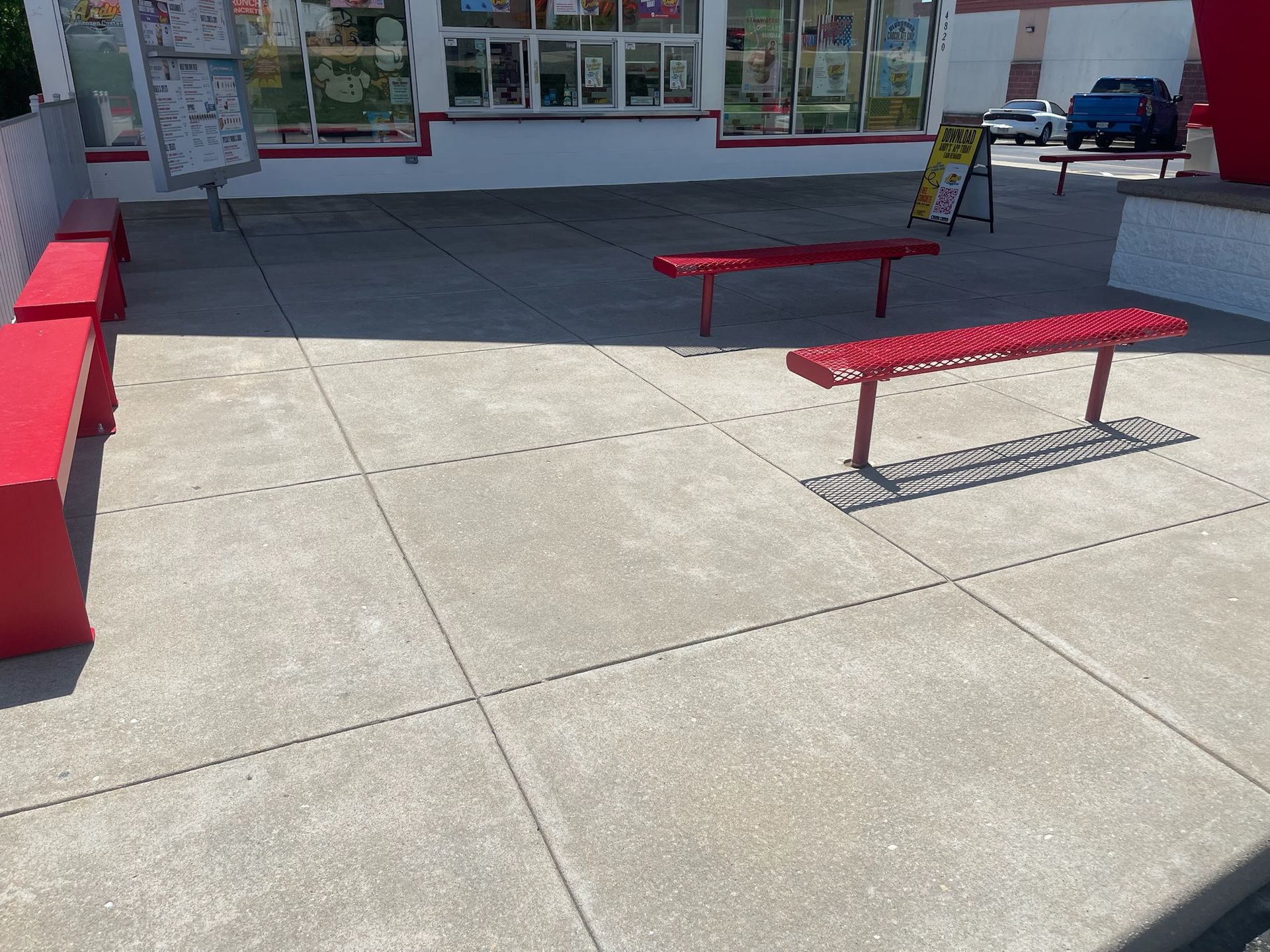Outdoor storefront patio with red benches on a concrete sidewalk in bright sunlight