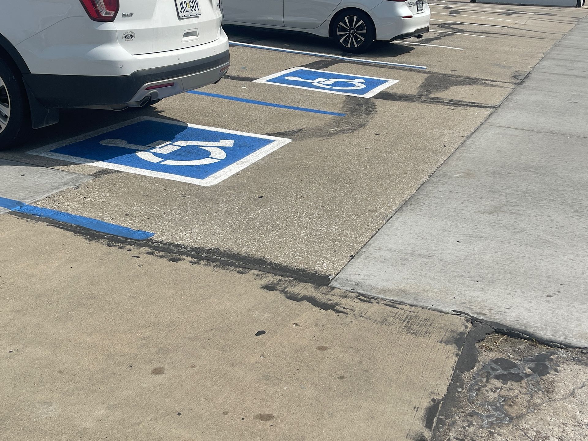 Accessible parking spaces on a cracked lot beside parked cars and a sidewalk