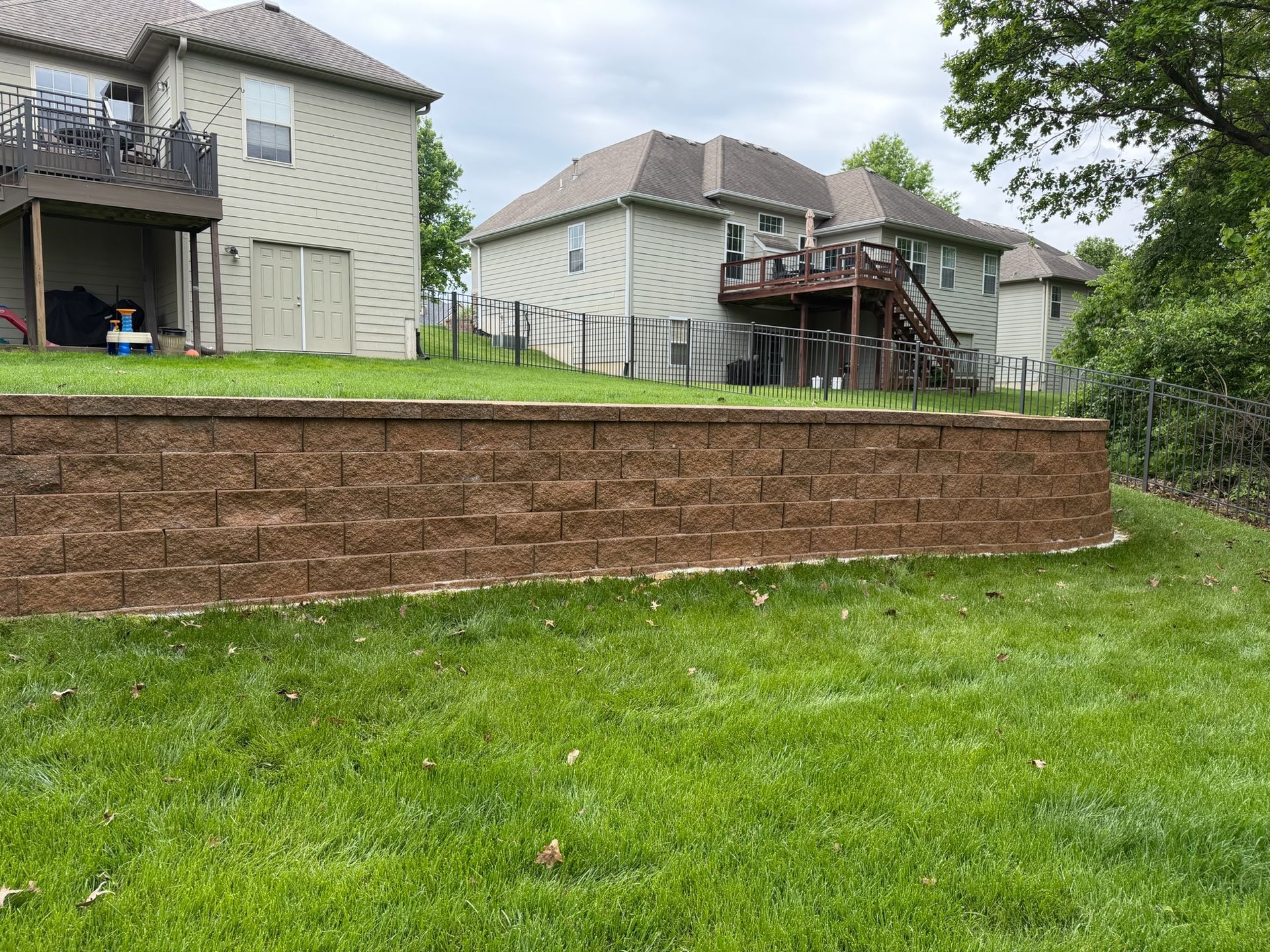 Suburban backyard with stone retaining wall, green lawn, and two tan houses on a cloudy day