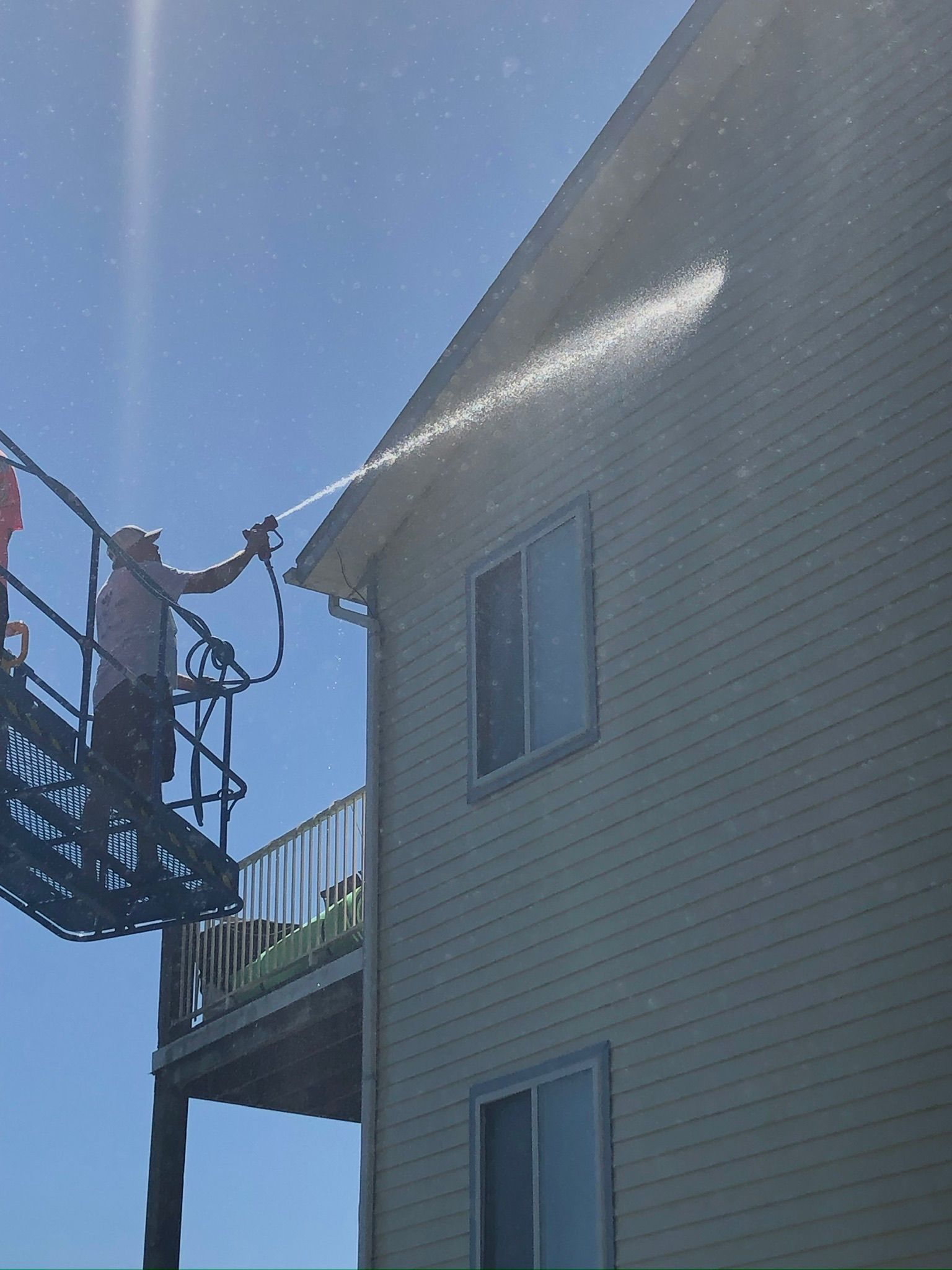 Person on a lift spraying the side of a beige house under a blue sky