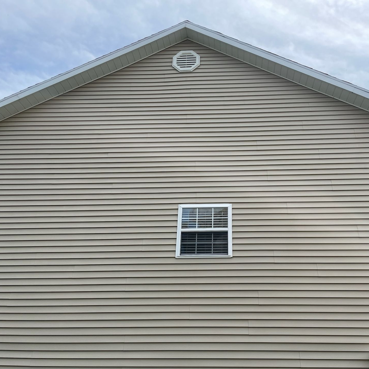 Beige house exterior with horizontal siding, centered window, and gable vent under a cloudy sky